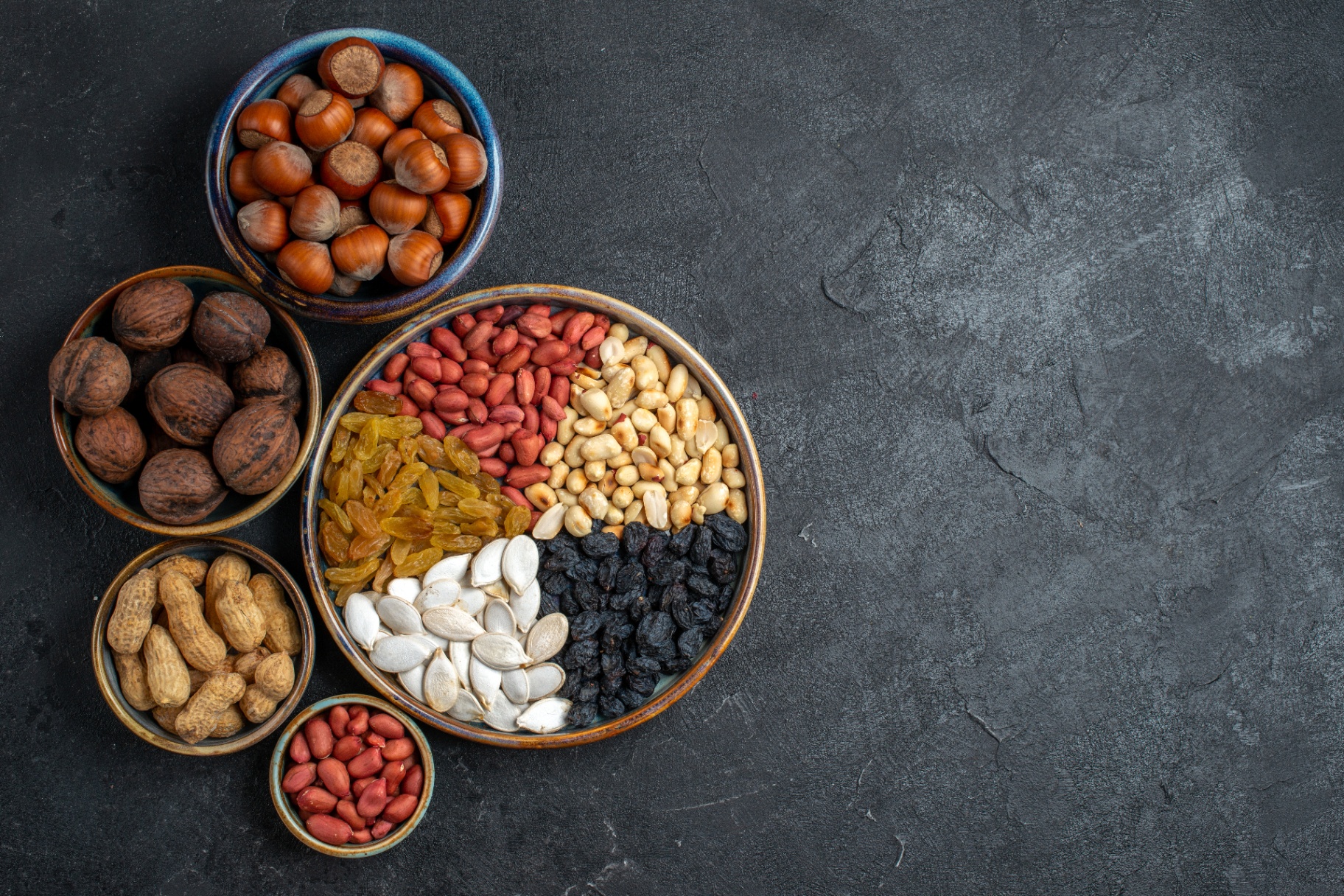 Top view of premium nuts, raisins and dried fruits in ceramic bowls on a dark slate background